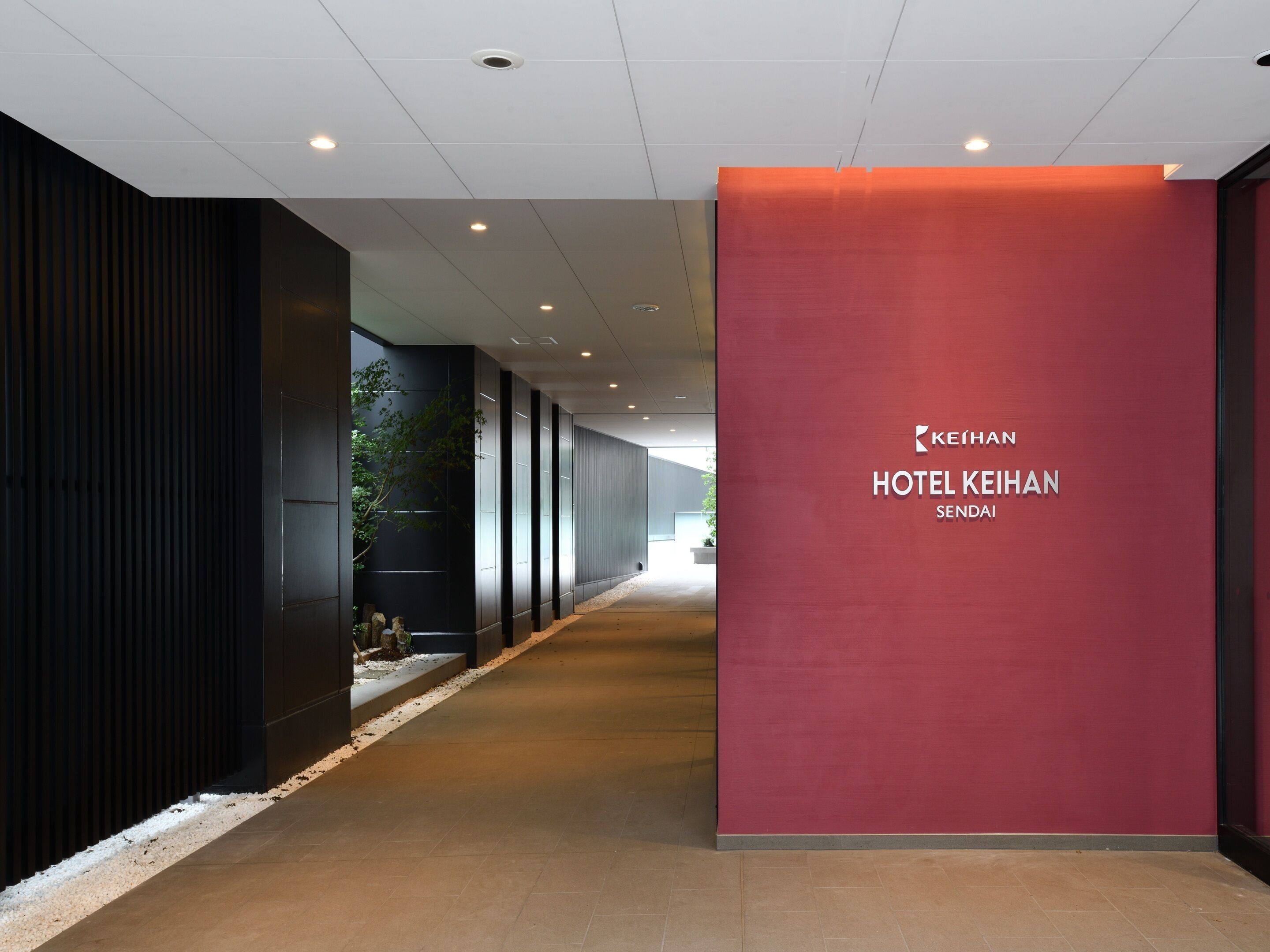 A hallway with a red wall displaying the hotel name, a black wall with vertical lines, and a tiled floor.
