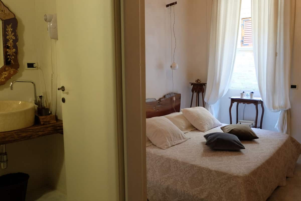 Une chambre à coucher comprenant un lit, une table de chevet, une petite table avec une lampe, une fenêtre avec des rideaux et une salle de bain avec un lavabo et un miroir.