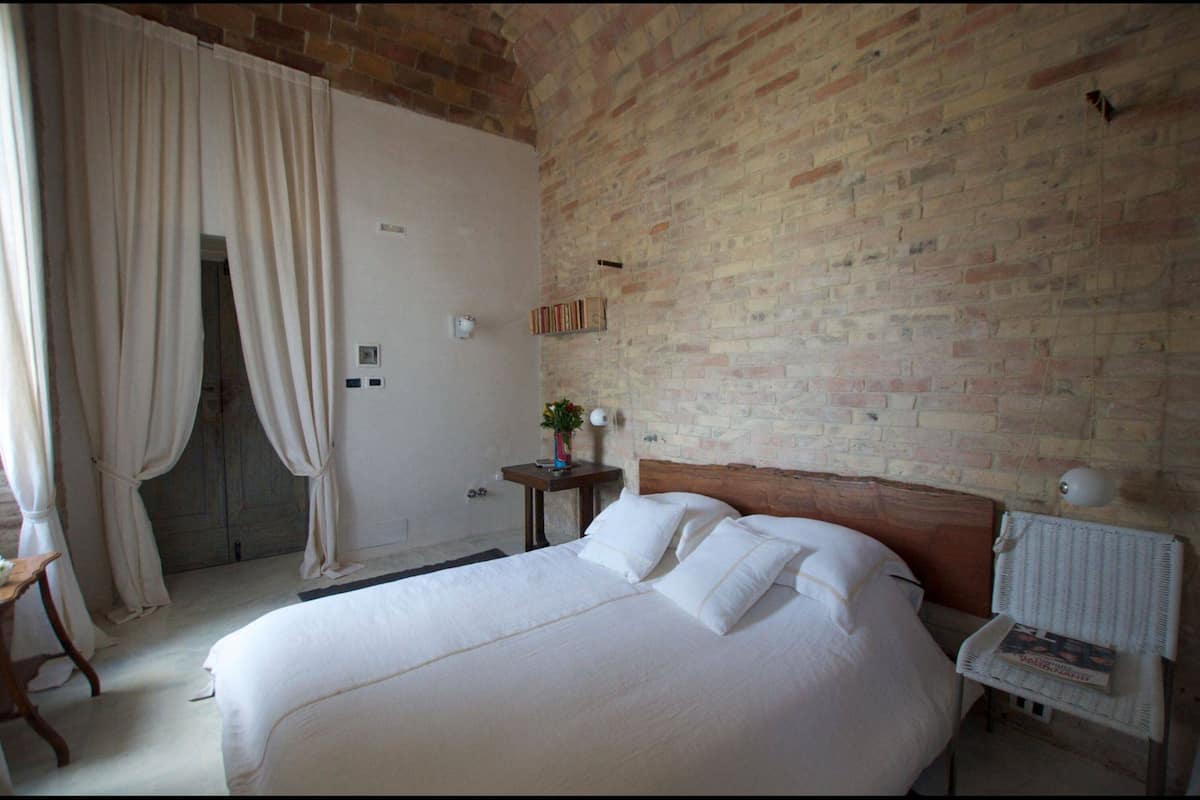 Une chambre avec un mur en briques, un cadre de lit en bois, une table de chevet avec une lampe et une porte recouverte d’un rideau.