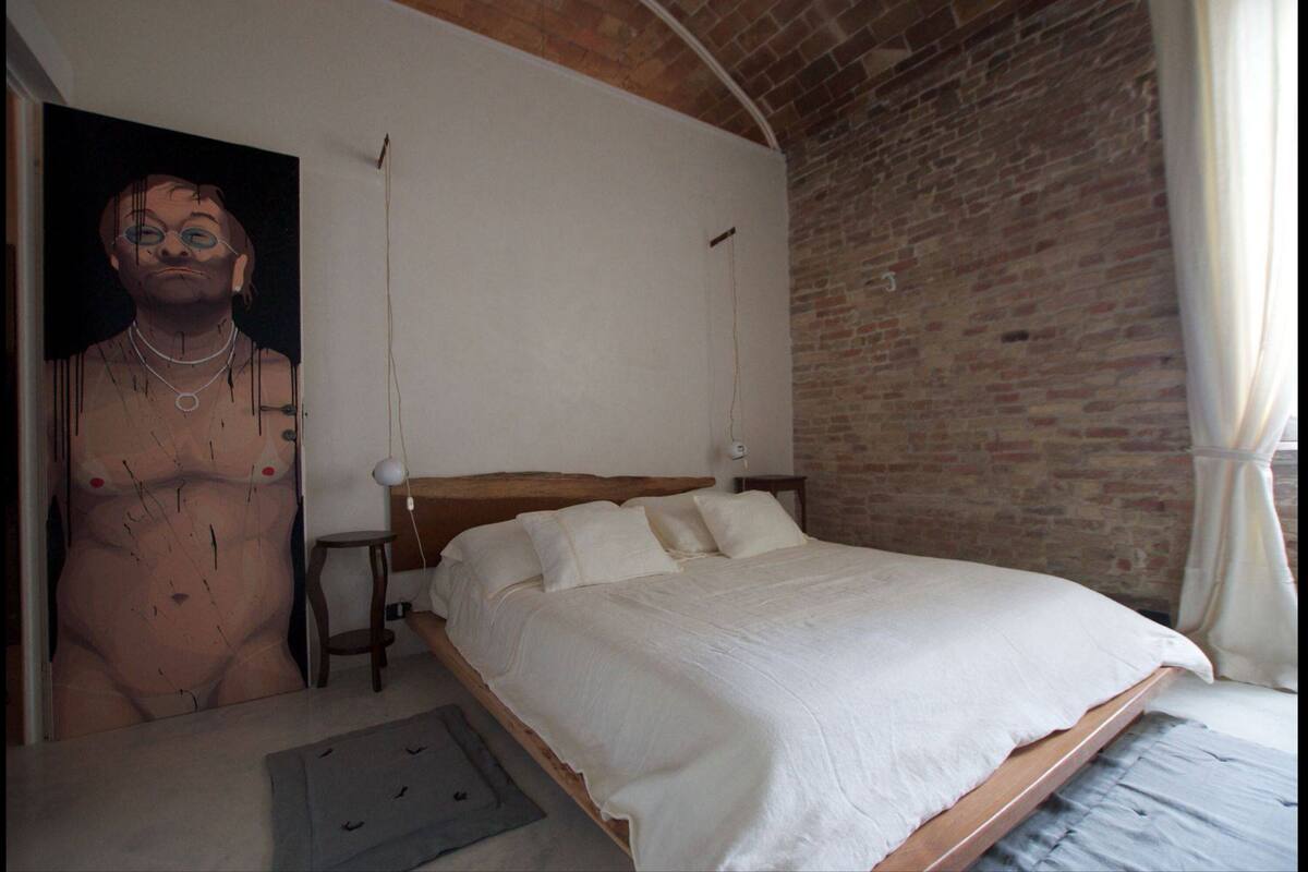 Une chambre avec un mur en briques, un cadre de lit en bois, du linge de lit blanc et un grand tableau accroché au mur.