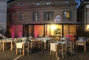 Un área de comedor al aire libre con mesas dispuestas para los invitados, fachada de un edificio de ladrillo y un gran ventanal iluminado.