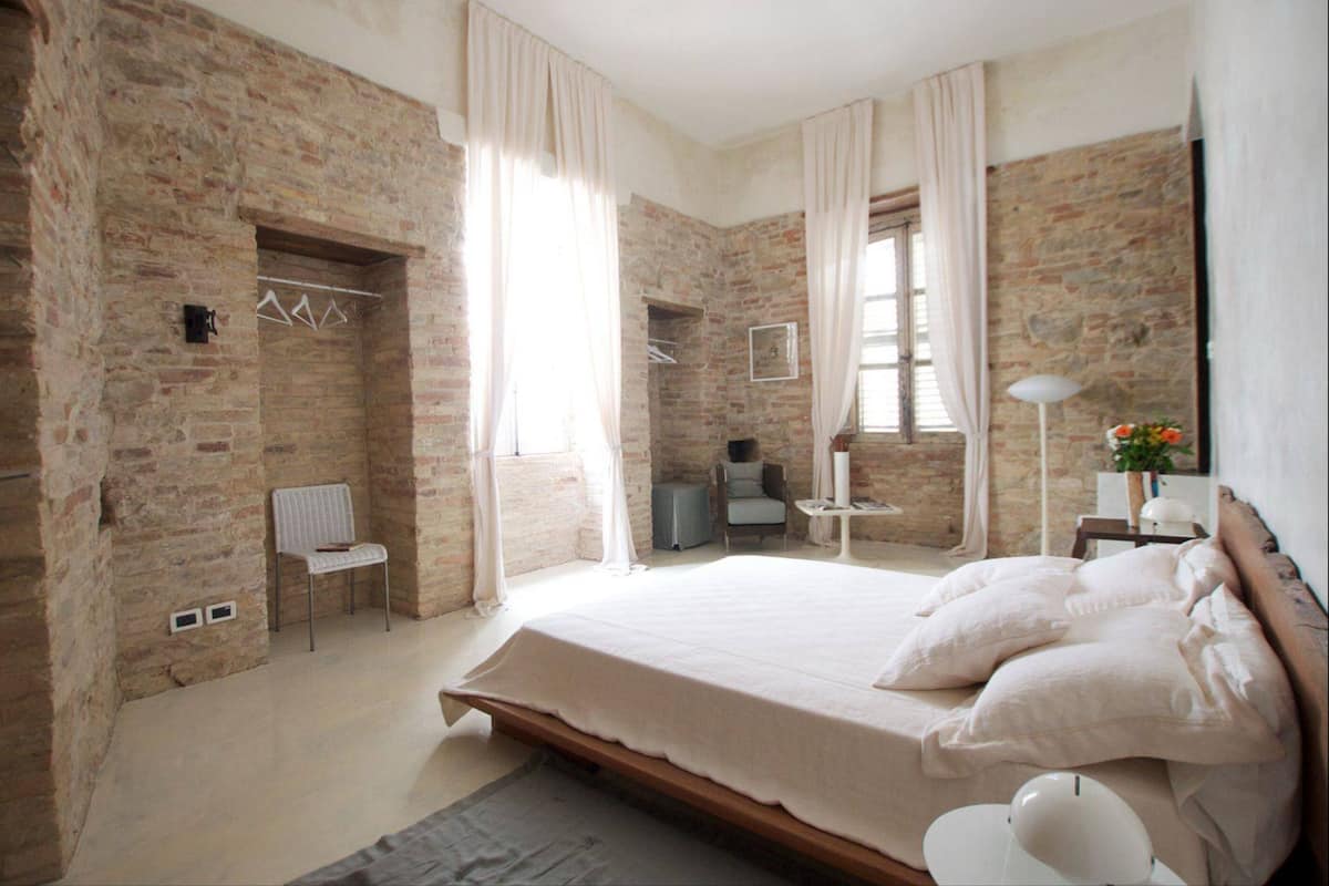 Une chambre avec des murs en briques apparentes, un grand lit avec du linge de lit blanc, une petite table avec une lampe et une chaise.