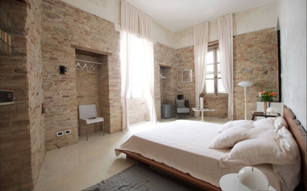 Un dormitorio con paredes de ladrillo visto, una cama grande con ropa blanca, una mesita con una lámpara y una silla.