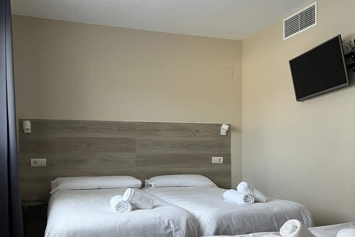 Habitación de hotel con tres camas, un televisor de pantalla plana montado en la pared y un cabecero de madera.