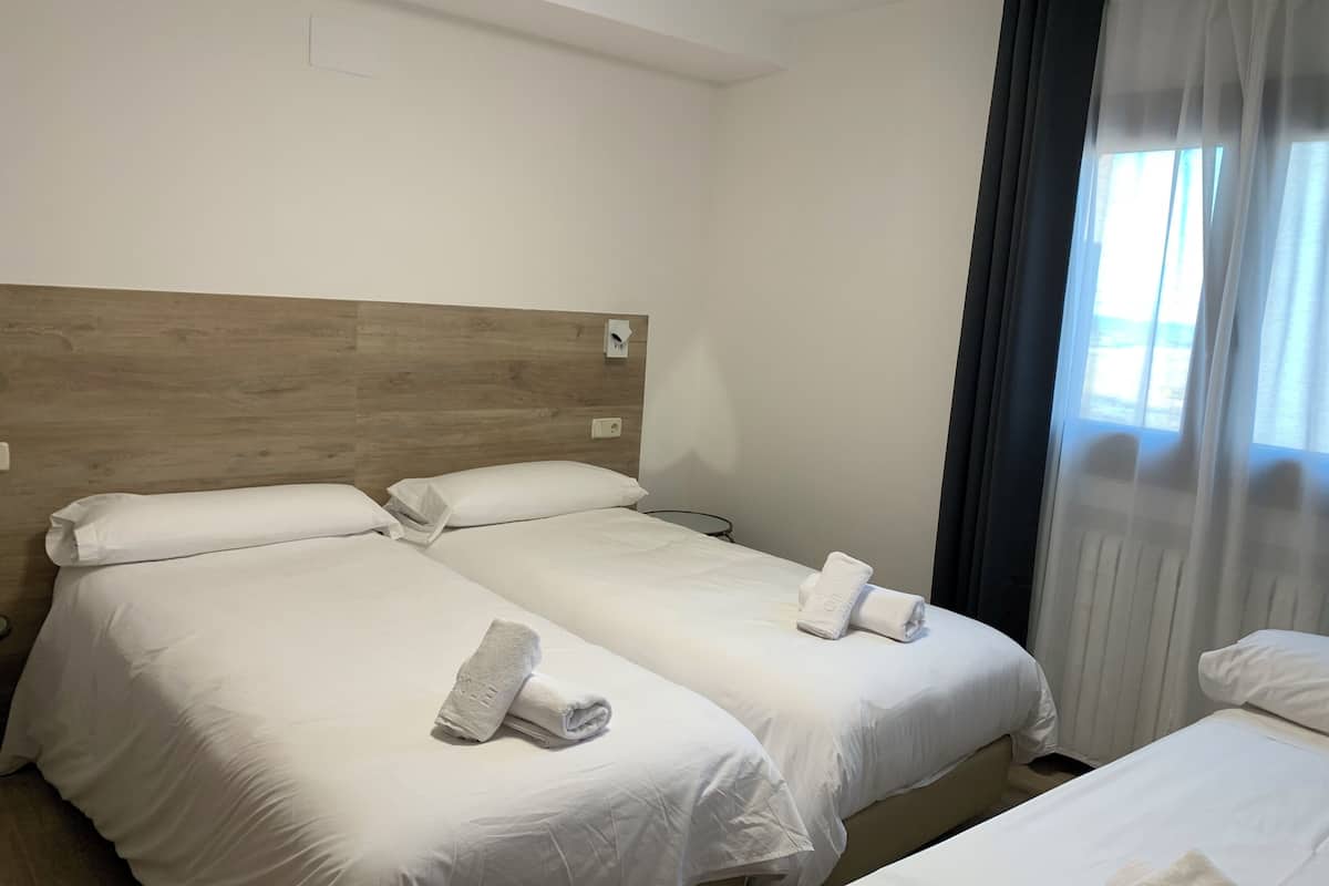 Habitación de hotel con dos camas individuales, cada una con ropa de cama y toallas blancas, cabeceros de madera, una ventana con cortinas y un enchufe en la pared.