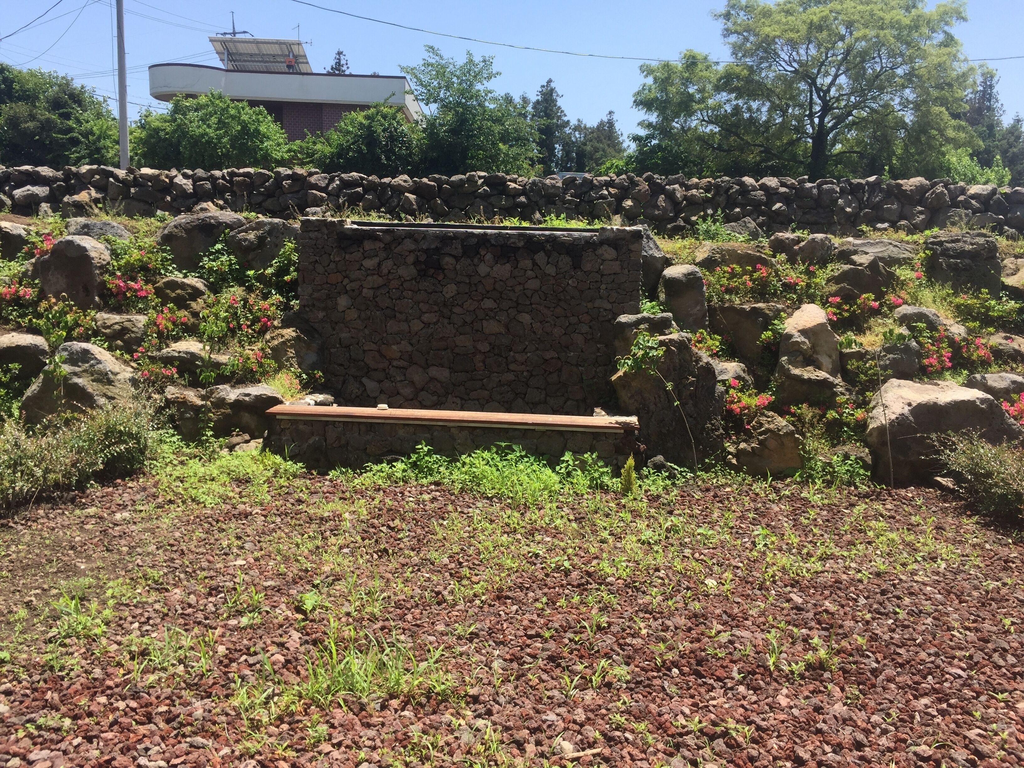 Eine Steinsitzbank, umgeben von Steinen und Grünpflanzen, mit einer Steinmauer und einem Gebäude im Hintergrund.