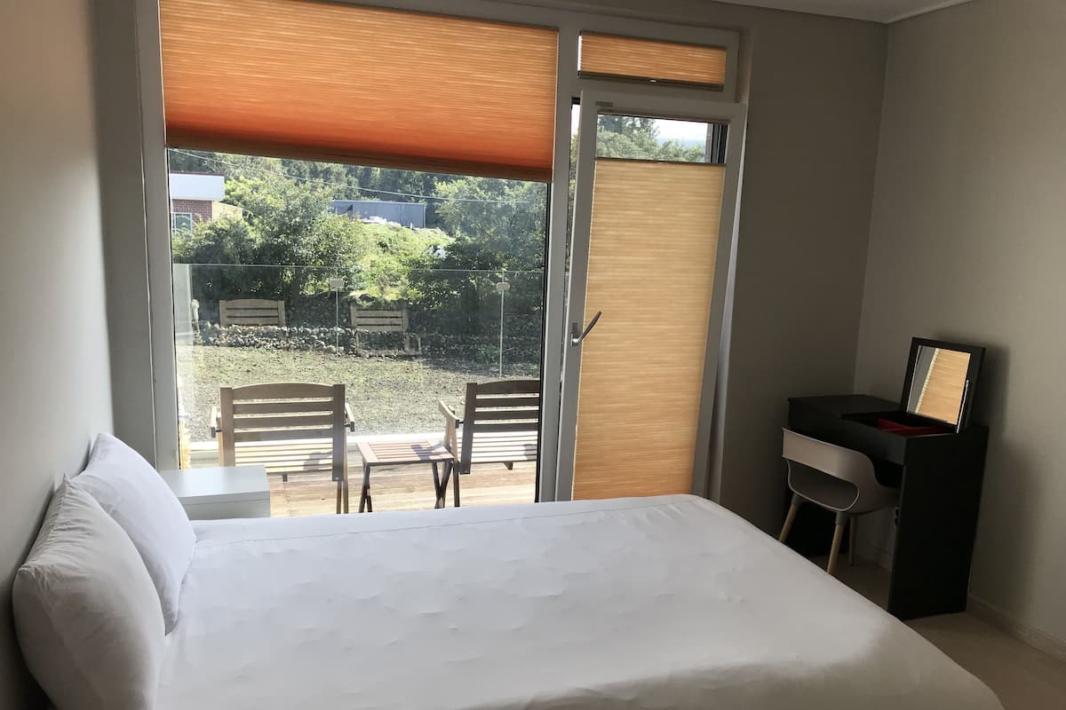 A bedroom with a bed, a desk, and a chair. There is a window with blinds, and a view of the outside.