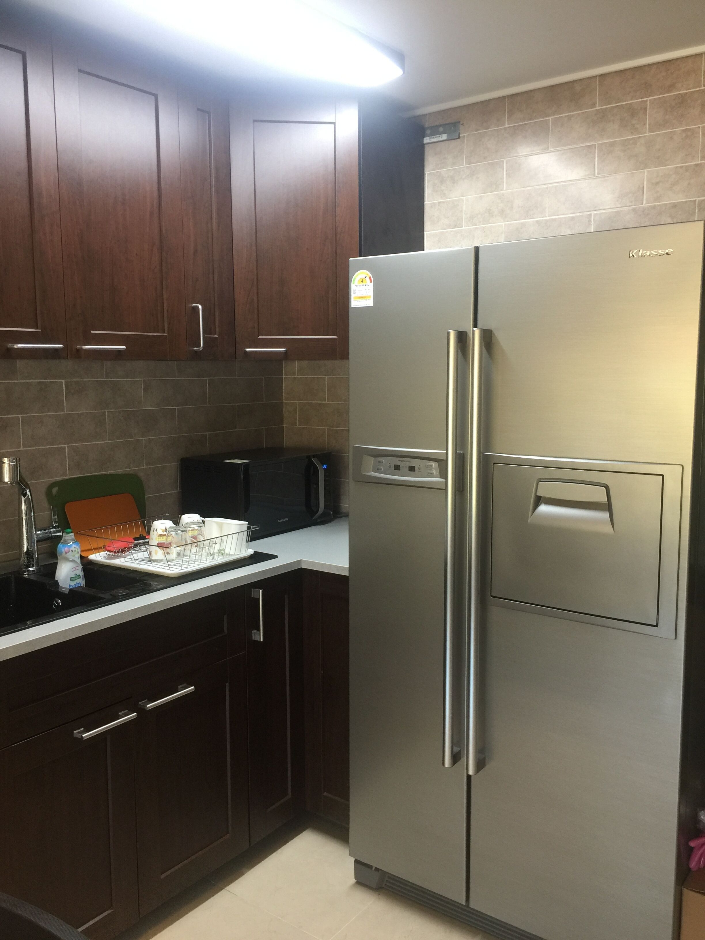 Una cocina con armarios de madera oscura, un refrigerador de acero inoxidable, un microondas negro y una encimera con un escurreplatos y una botella de leche.