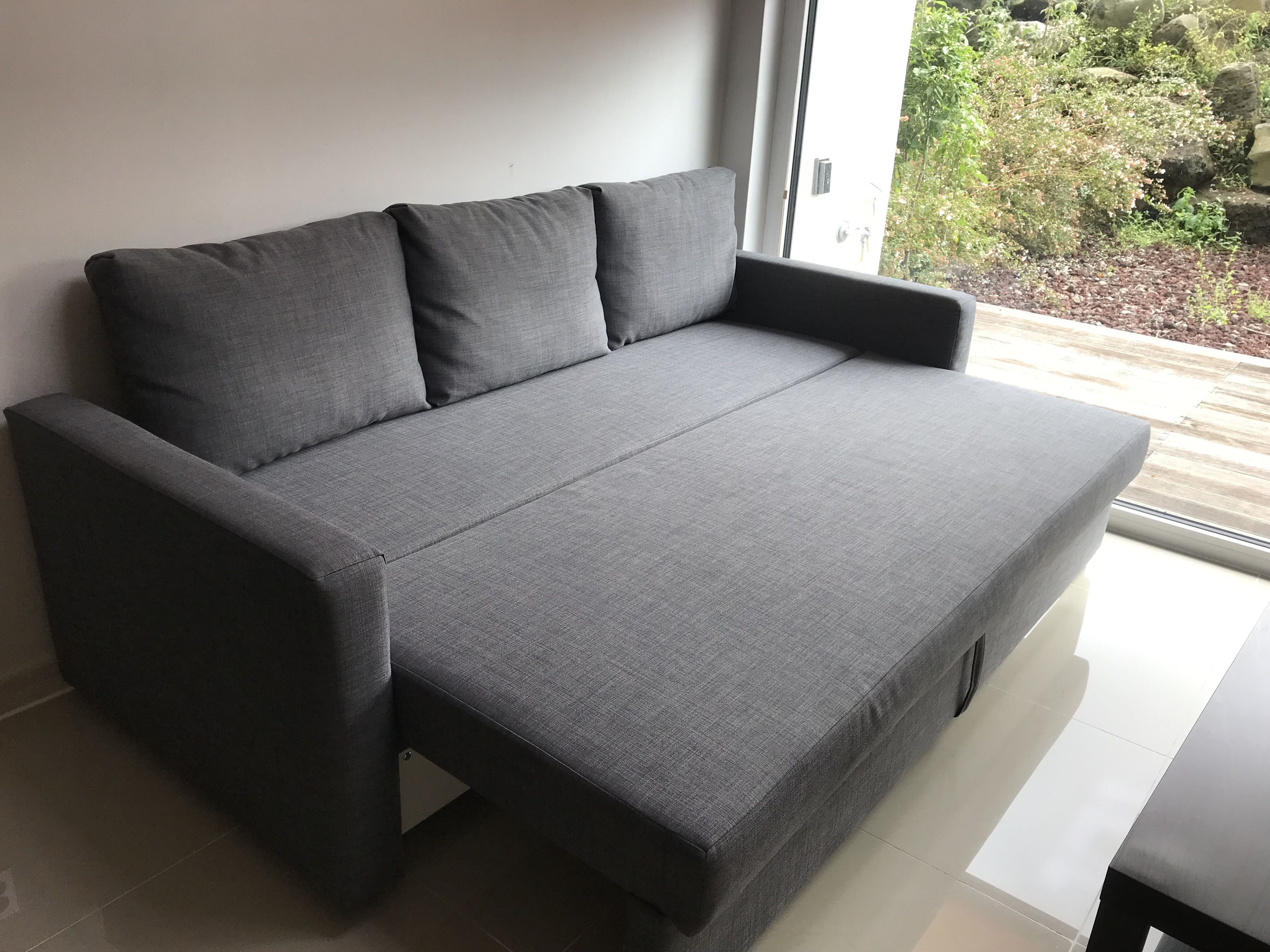A grey sofa with a low profile and a pull-out section, positioned against a white wall with a window that offers a view of greenery outside.