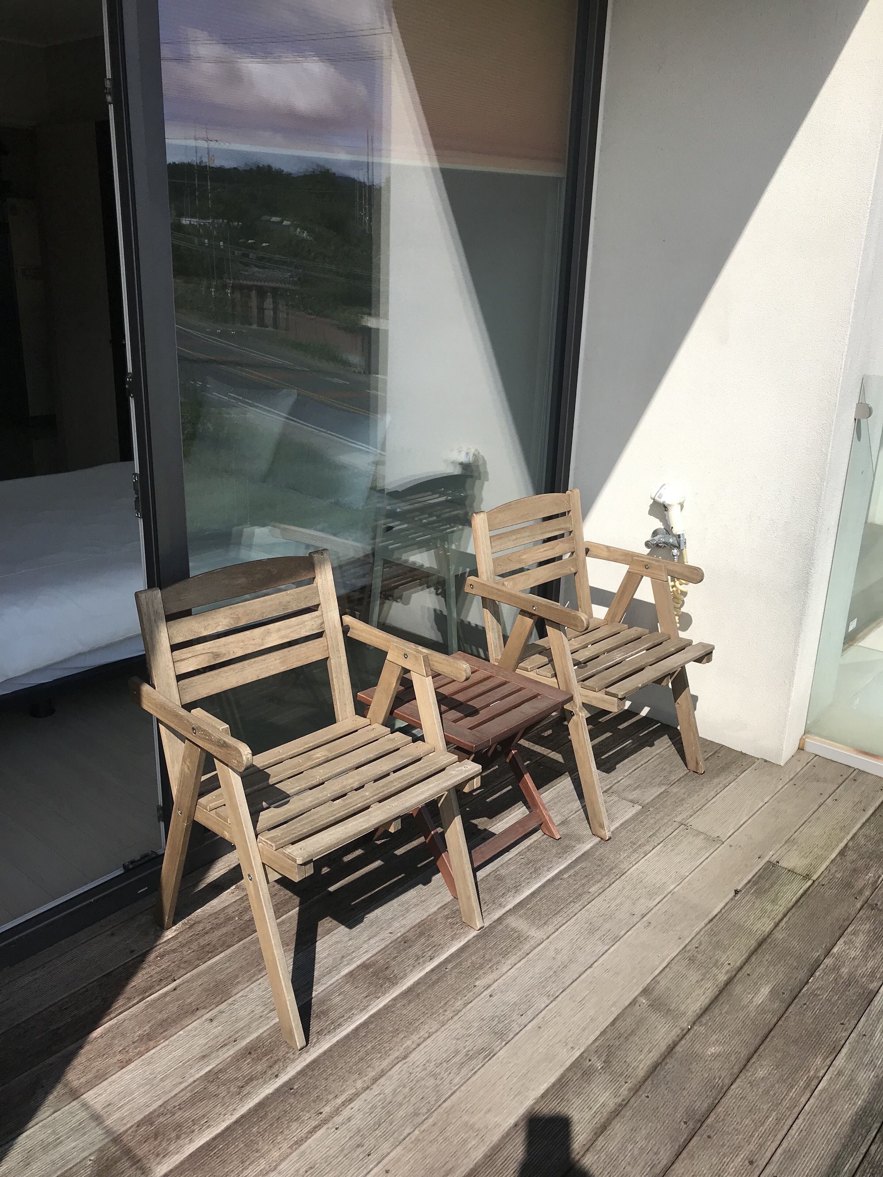 Eine hölzerne Bank auf einer Holzterrasse mit einer Glastür, die einen Blick auf eine Straße und Grünflächen bietet.