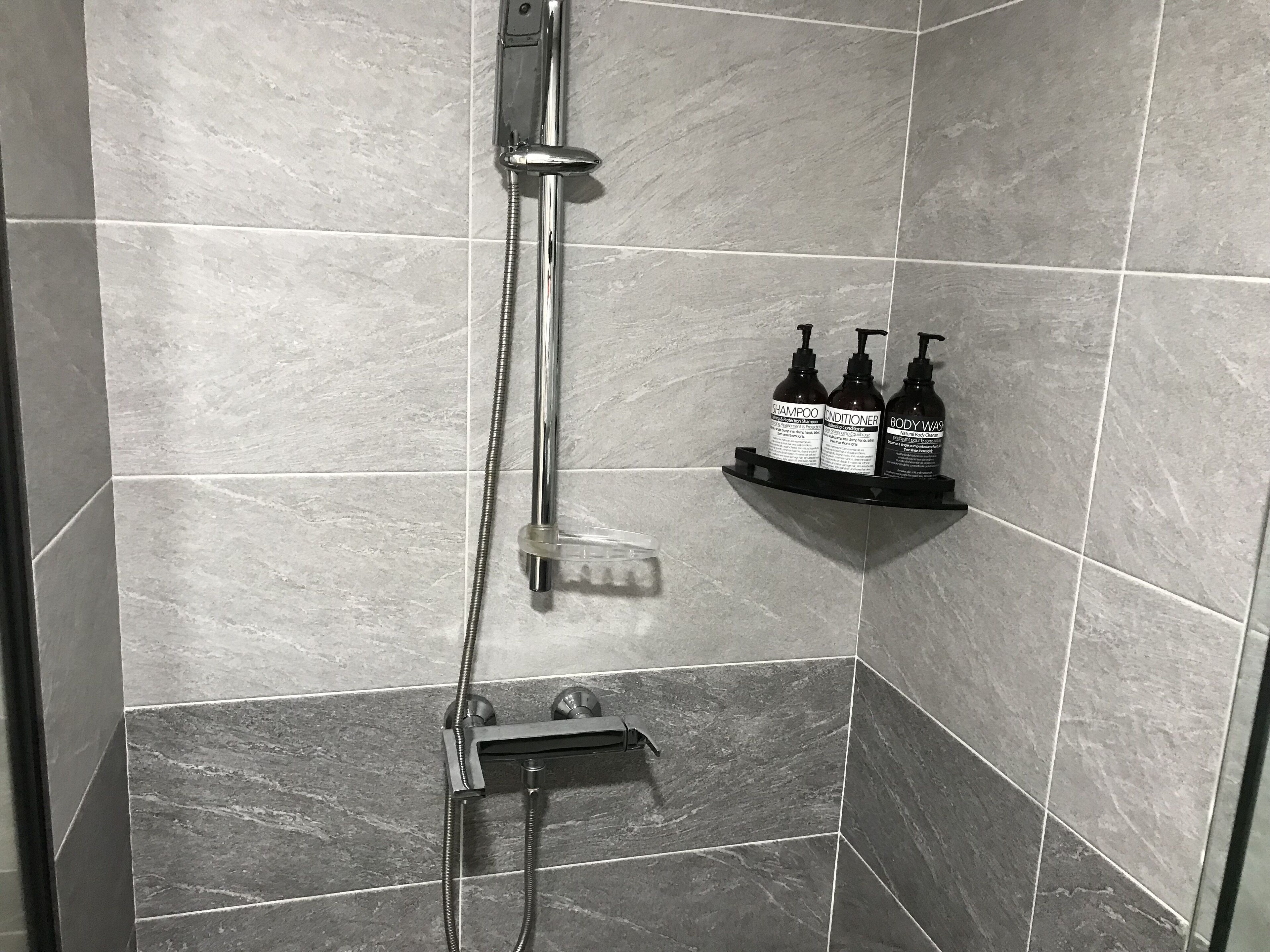A shower area with a handheld showerhead, a wall-mounted faucet, and a shelf with three bottles of shampoo, conditioner, and body wash.