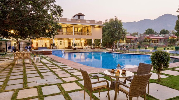 A poolside area with a dining table and chairs, a pool, and a two-story building in the background. - Vatsalya Vihar - A Luxury Pool Villas Resort (Udaipur)