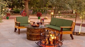 An outdoor seating area with two green cushioned chairs, a round wooden table, and a metal fire pit with a visible fire. - Vatsalya Vihar - A Luxury Pool Villas Resort (Udaipur)