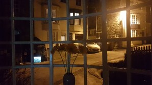 Een raam met uitzicht op een gebouw 's nachts, een vaas met bloemen, en een geparkeerde auto buiten.