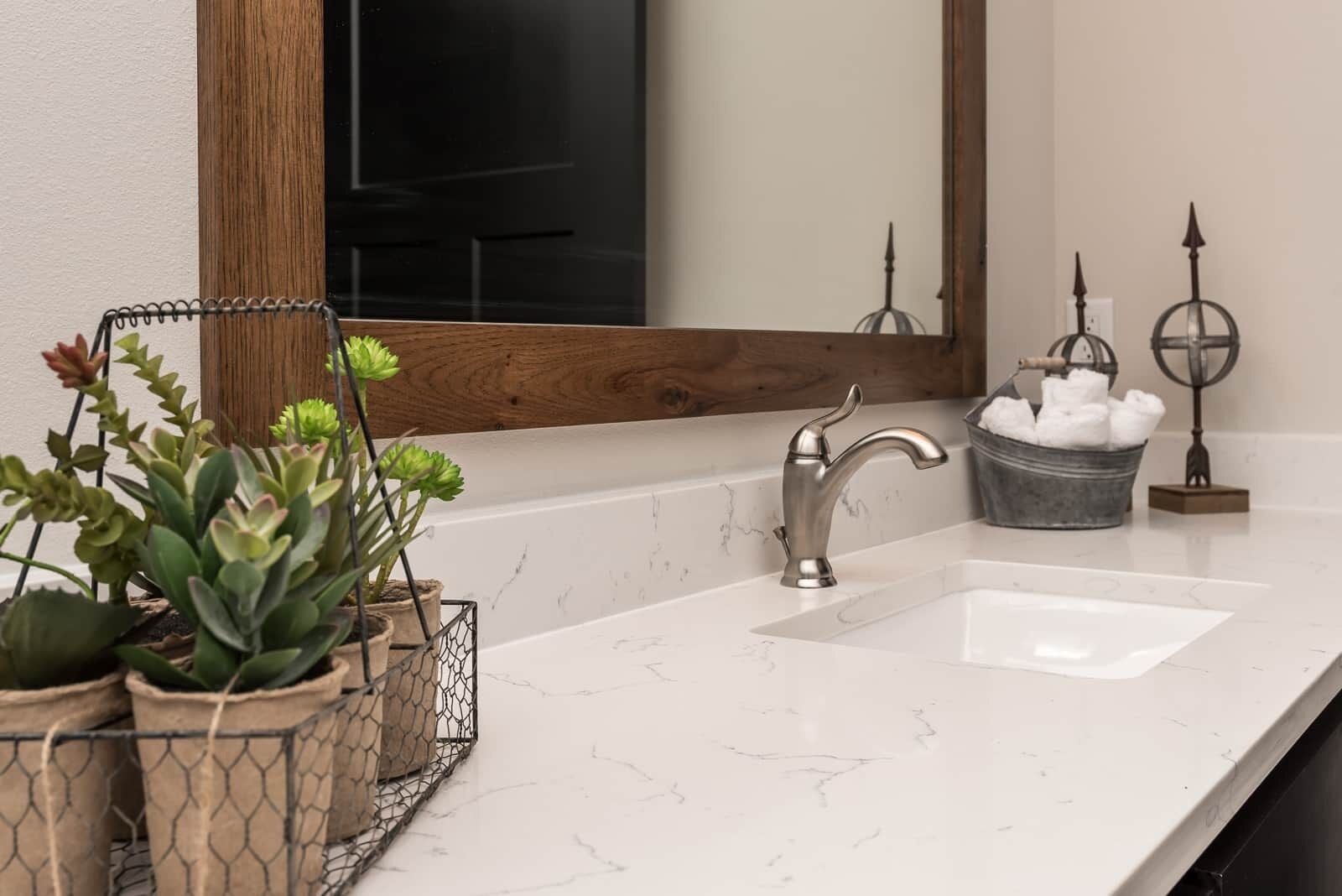 A bathroom with a white marble sink, a modern faucet, a wooden-framed mirror, and potted plants.