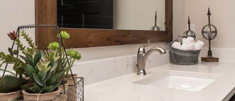 A bathroom with a white marble sink, a modern faucet, a wooden-framed mirror, and potted plants.