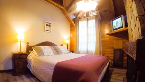Een knus slaapkamer met een bed, twee bedlampjes, een televisie op een plank, en een raam met gordijnen.