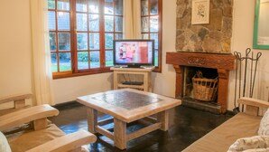 Een gezellige kamer met een stenen openhaard, een televisie op een houten dressoir, en uitzicht op de tuin door grote ramen.