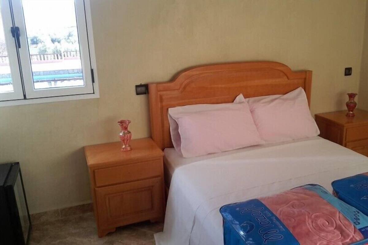 A bedroom with a wooden headboard, a bed with white bedding, two bedside tables with pink lamps, and a window with a view of a building.