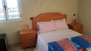 A bedroom with a wooden headboard, a bed with white bedding, two bedside tables with pink lamps, and a window with a view of a building.