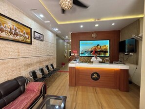 Un espace d’accueil d’hôtel comprenant un comptoir de réception, un téléviseur diffusant des images de paysages, une horloge et un coin salon avec des fauteuils.