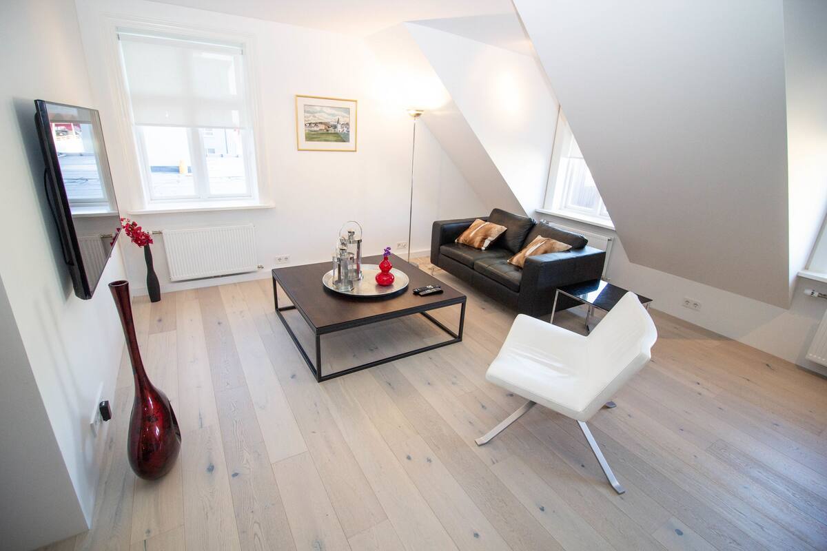 A modern loft apartment with a high ceiling, a black leather sofa, a white chair, a coffee table, and a red vase.