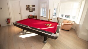 A pool table in a room with wooden flooring, a fire extinguisher, a window with blinds, and a framed picture on the wall. - City Point Reykjavik Center (Reykjavik)