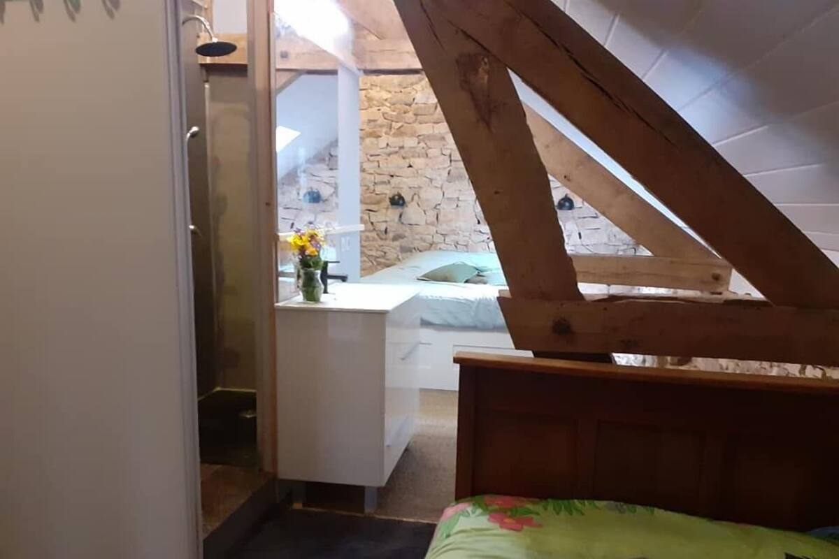 Une chambre avec un plafond en pente, une tête de lit en bois, un lit recouvert d’une couverture colorée, un bureau agrémenté d’un vase de fleurs et, au fond, un mur en pierre.