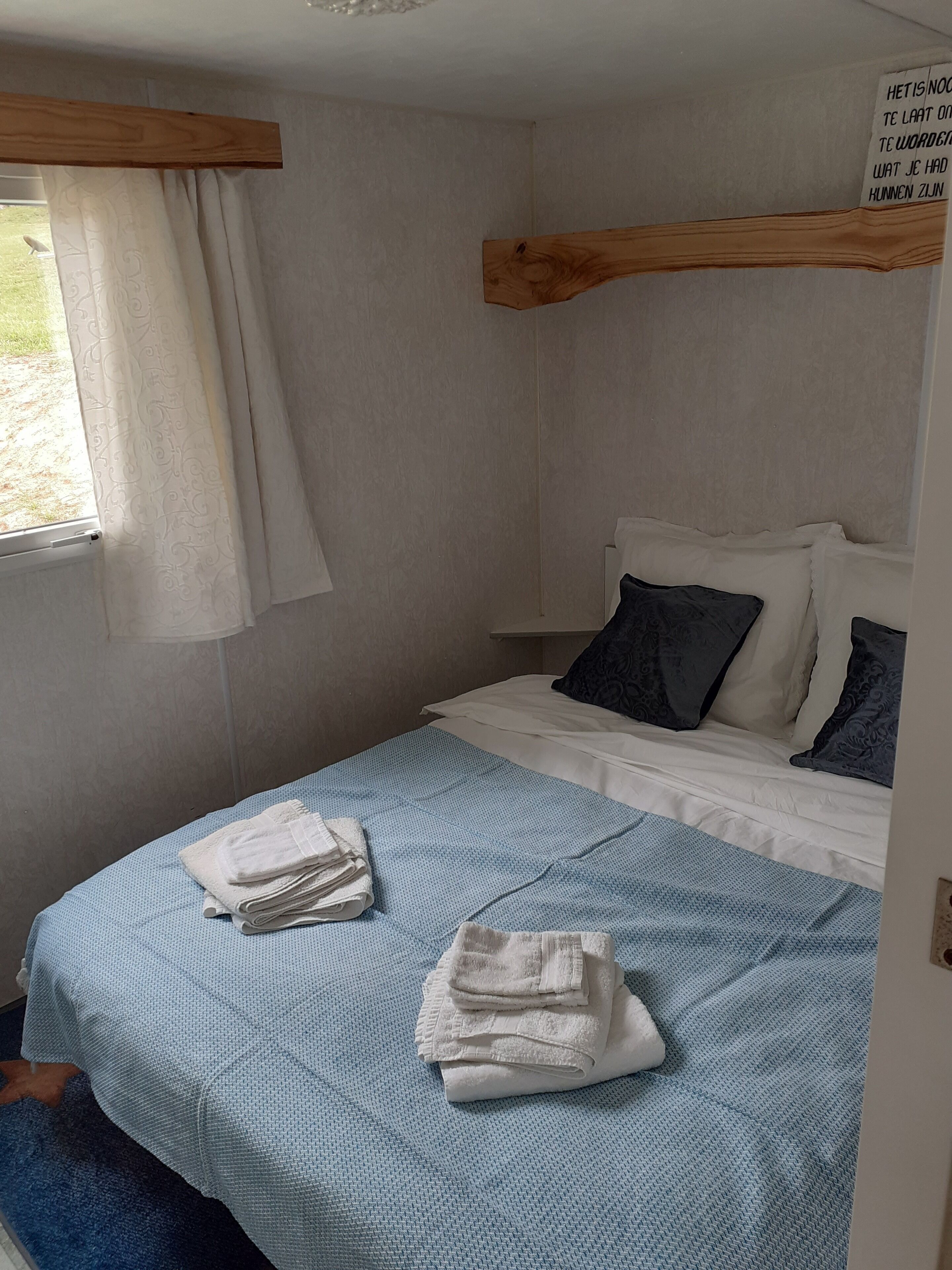 Une petite chambre avec un lit simple, une couvre-lit bleue, des oreillers blancs et une tête de lit en bois. Il y a une fenêtre avec un rideau blanc, et des serviettes sont soigneusement pliées sur le lit.