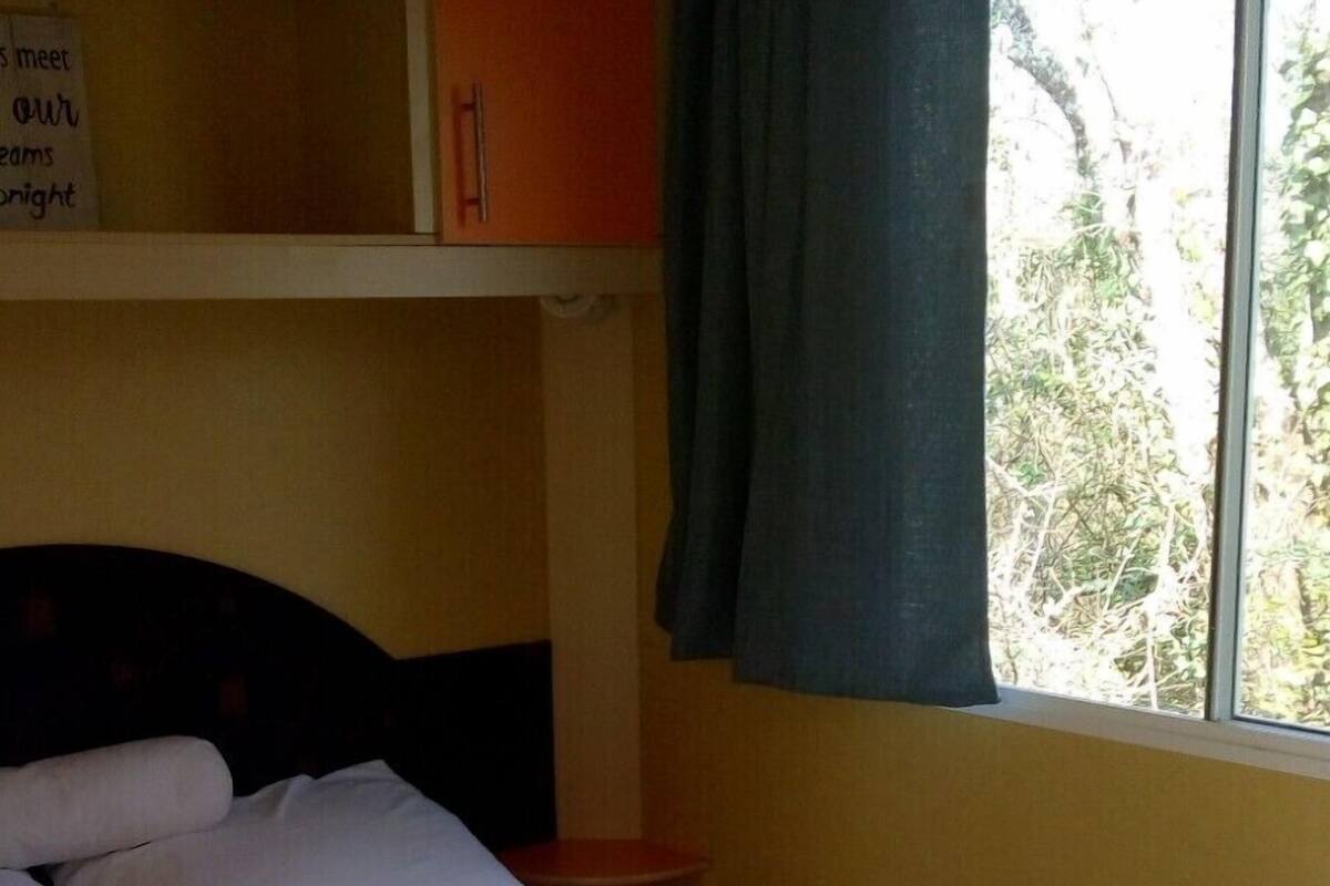 Un lit simple avec une couette blanche, dans une chambre dotée d’une tête de lit en bois, d’une fenêtre avec un rideau et d’une étagère sur laquelle est posé un panneau.