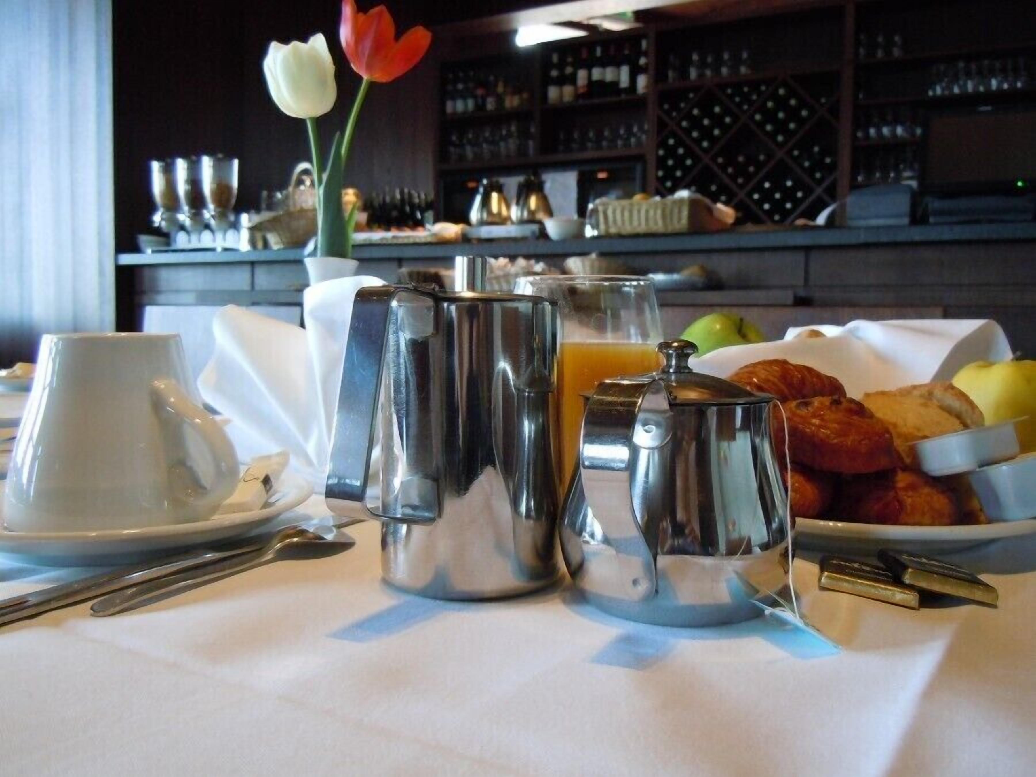 A breakfast setup with a coffee pot, teapot, and cups on a table, accompanied by croissants, apples, and a bowl of fruit.