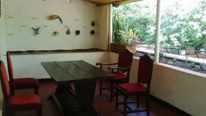 Un porche cubierto con una mesa de madera y sillas rojas con cojines, una pared blanca decorada con conchas marinas, y vistas de un jardín con plantas y flores.