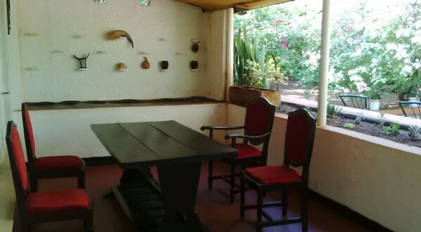 A covered patio with a wooden table and red cushioned chairs, a white wall with decorative shells, and a view of a garden with plants and flowers.