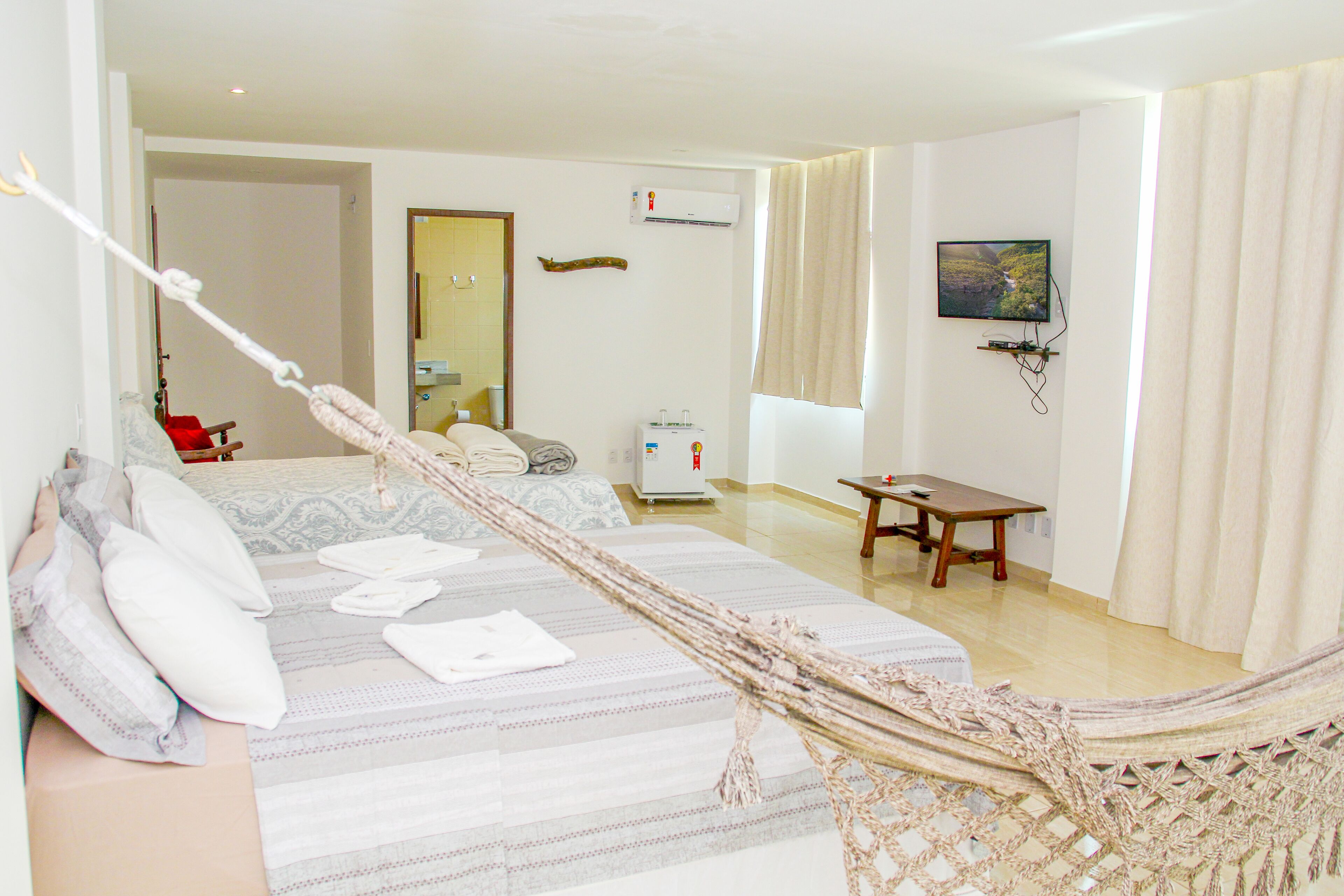 A hotel room with a bed, a hammock, a desk, a chair, a television, and a window with curtains.