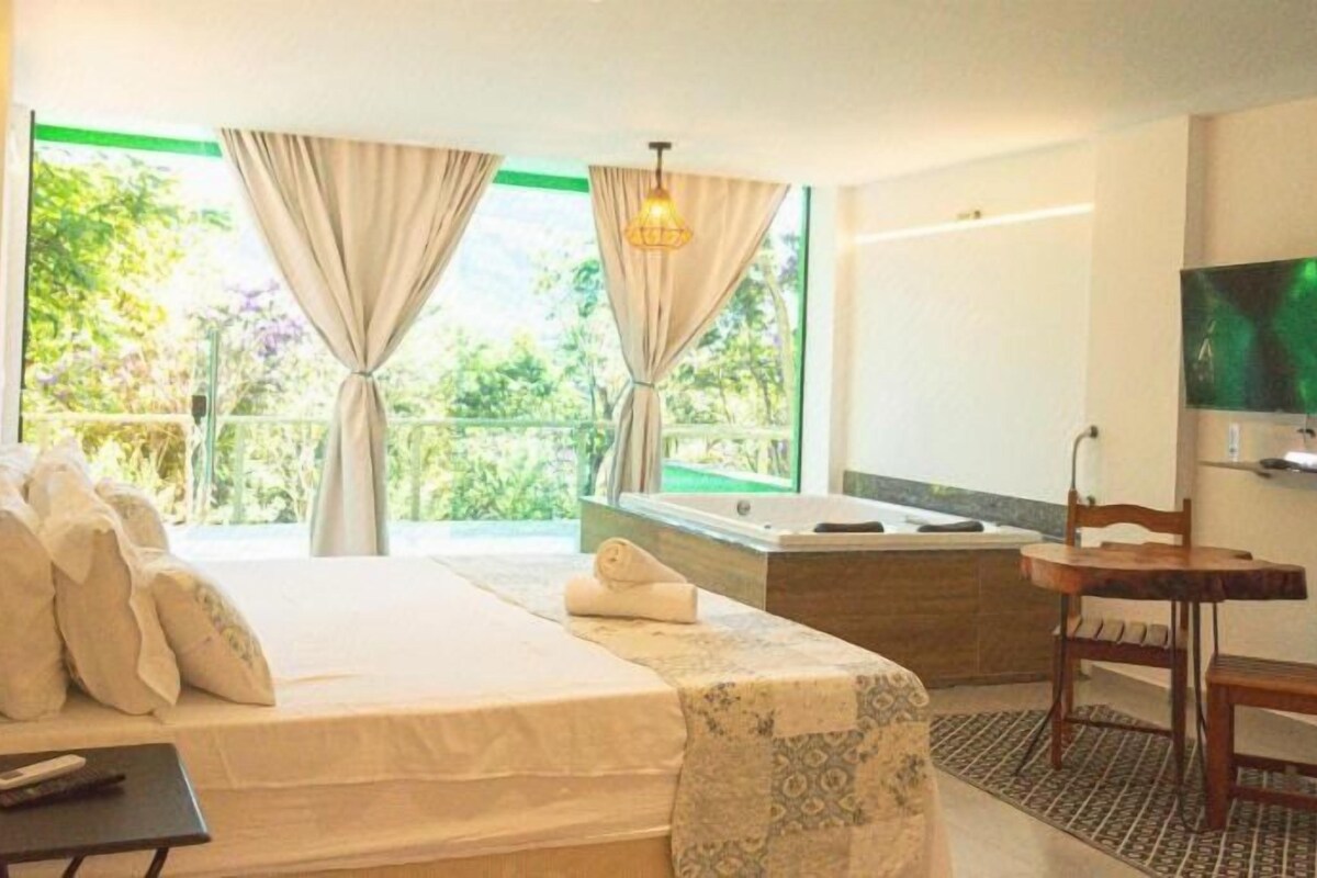 A hotel room with a bed, a desk, a TV, a bathtub, and a view of greenery.