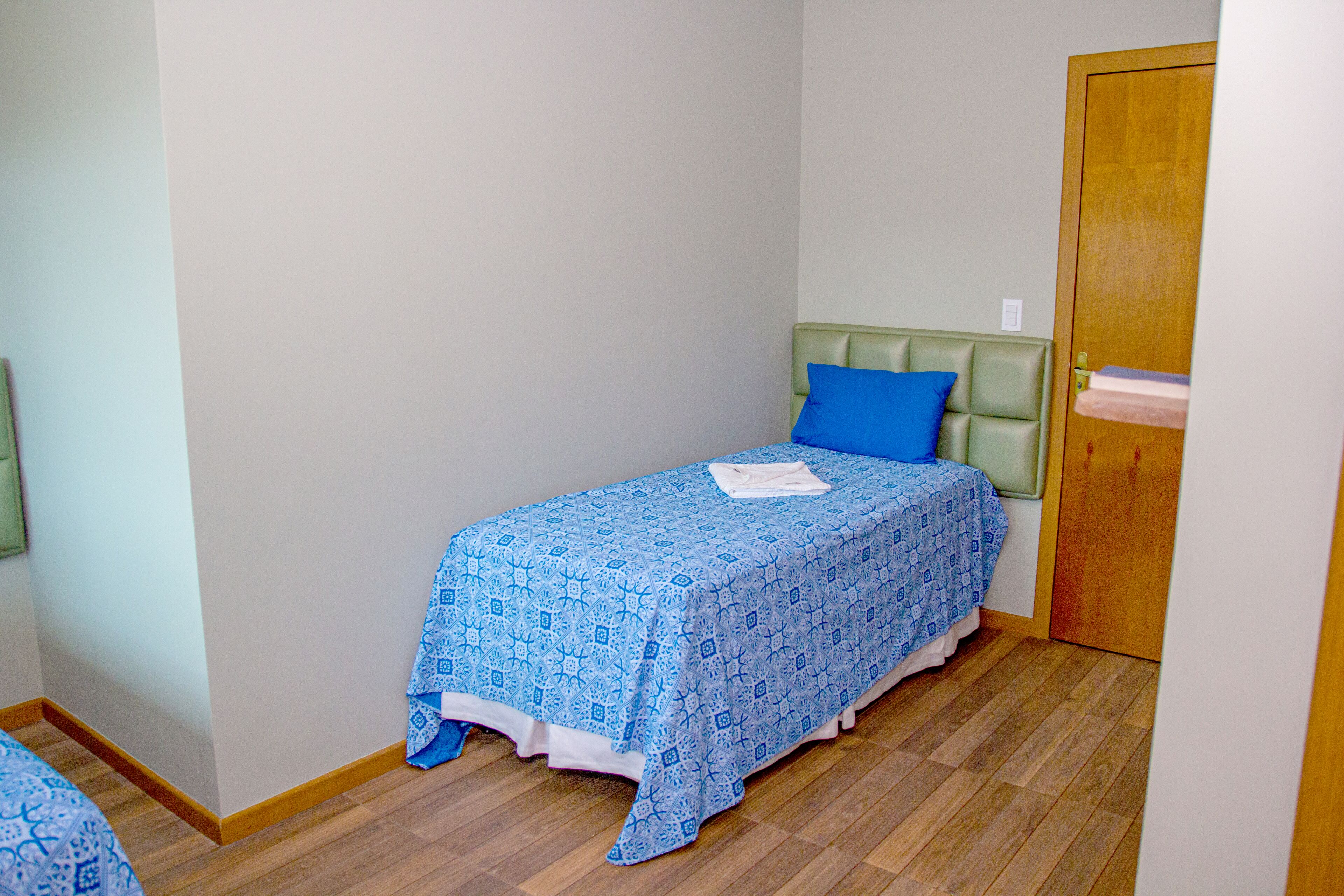 Una habitación pequeña con una cama individual, cabecera verde, colcha estampada en azul y puerta de madera.