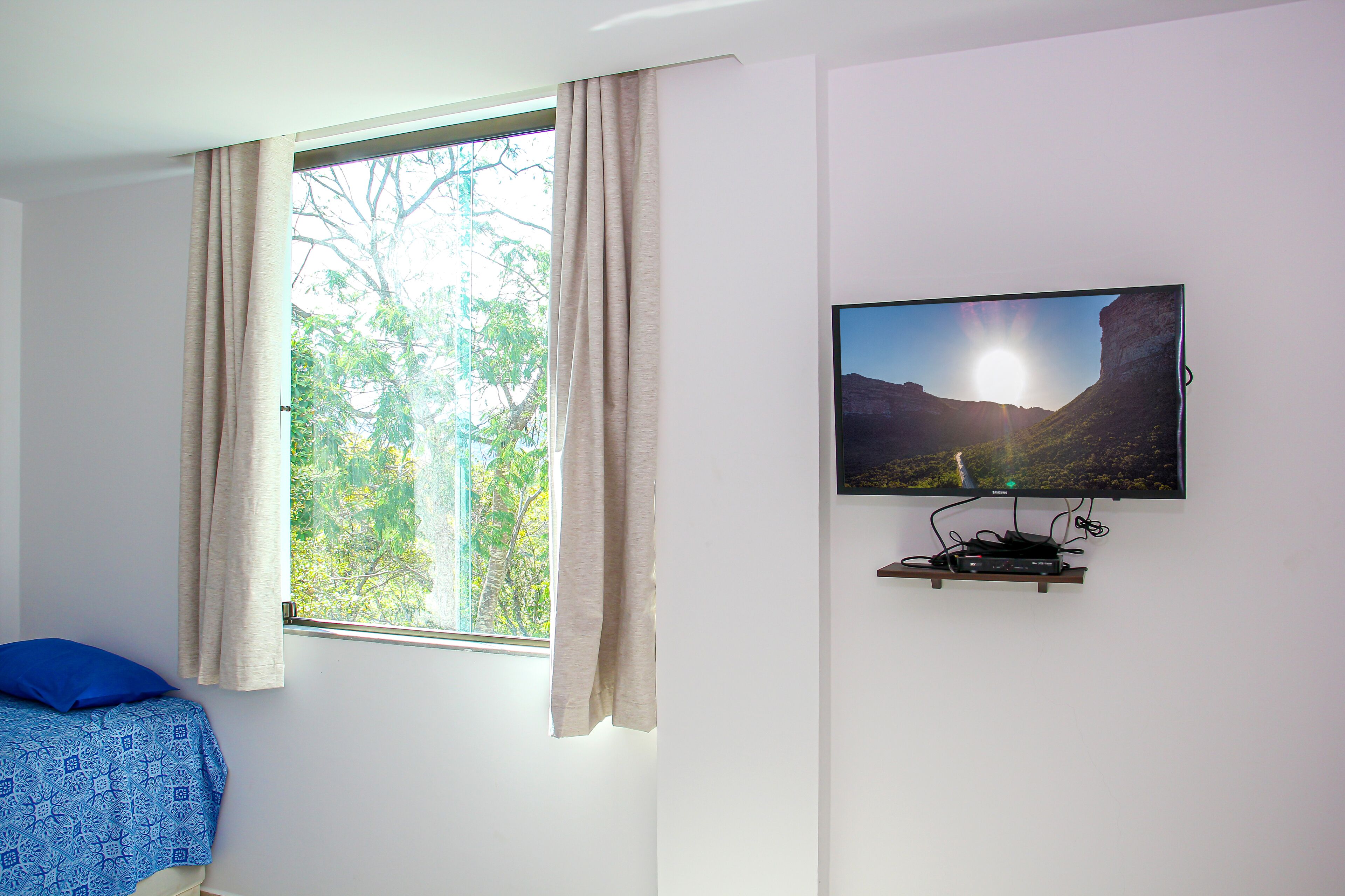 A bedroom with a bed, a window with curtains, and a wall-mounted TV displaying a landscape.