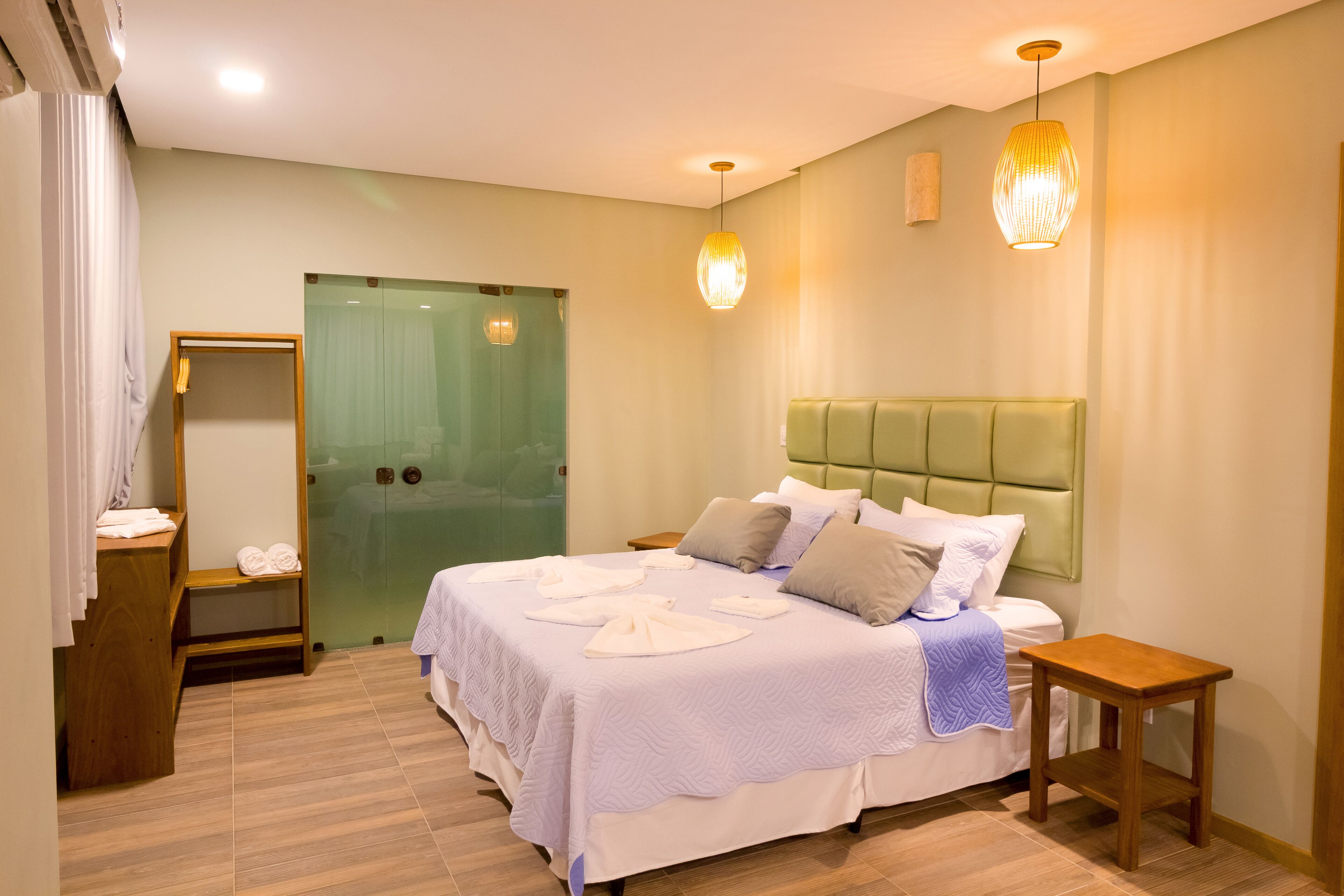 Habitación de hotel con cama, mesitas de noche, espejo, cabecero tapizado de color verde y dos lámparas colgantes.