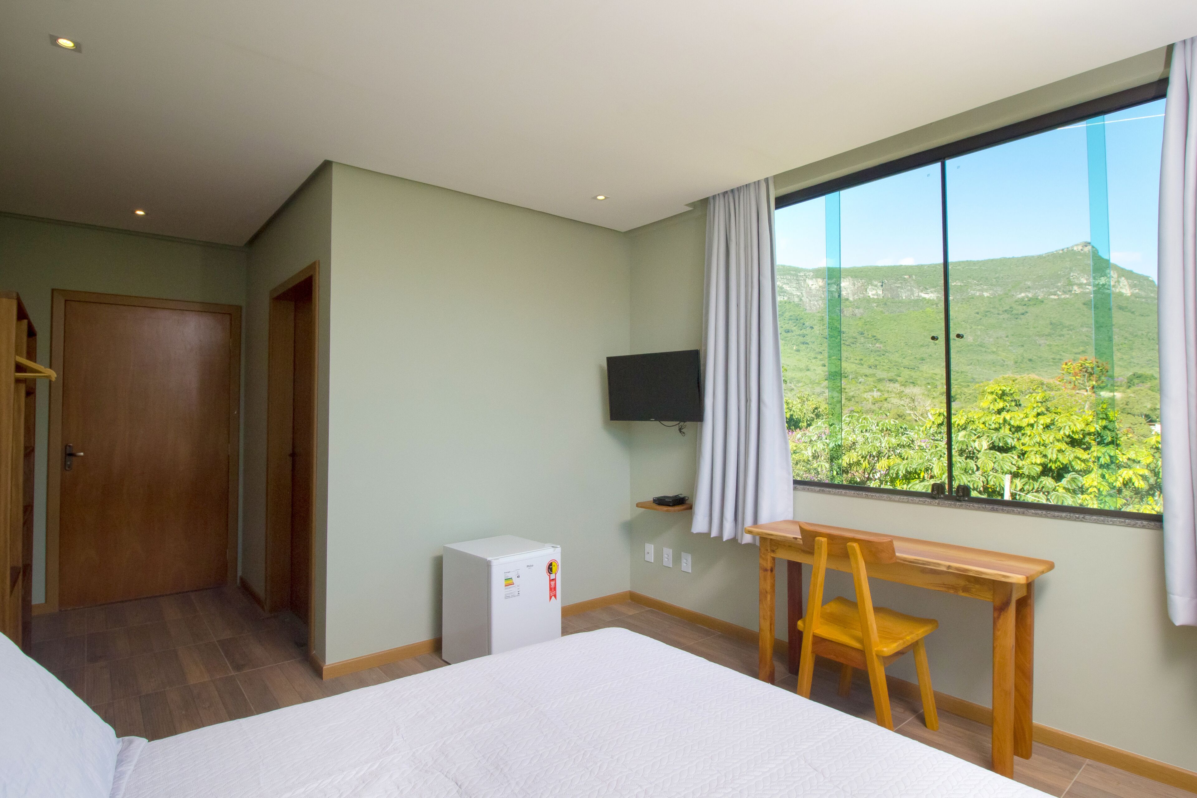 A hotel room with a bed, a small desk with a chair, a television, and a window with a view of greenery and hills.
