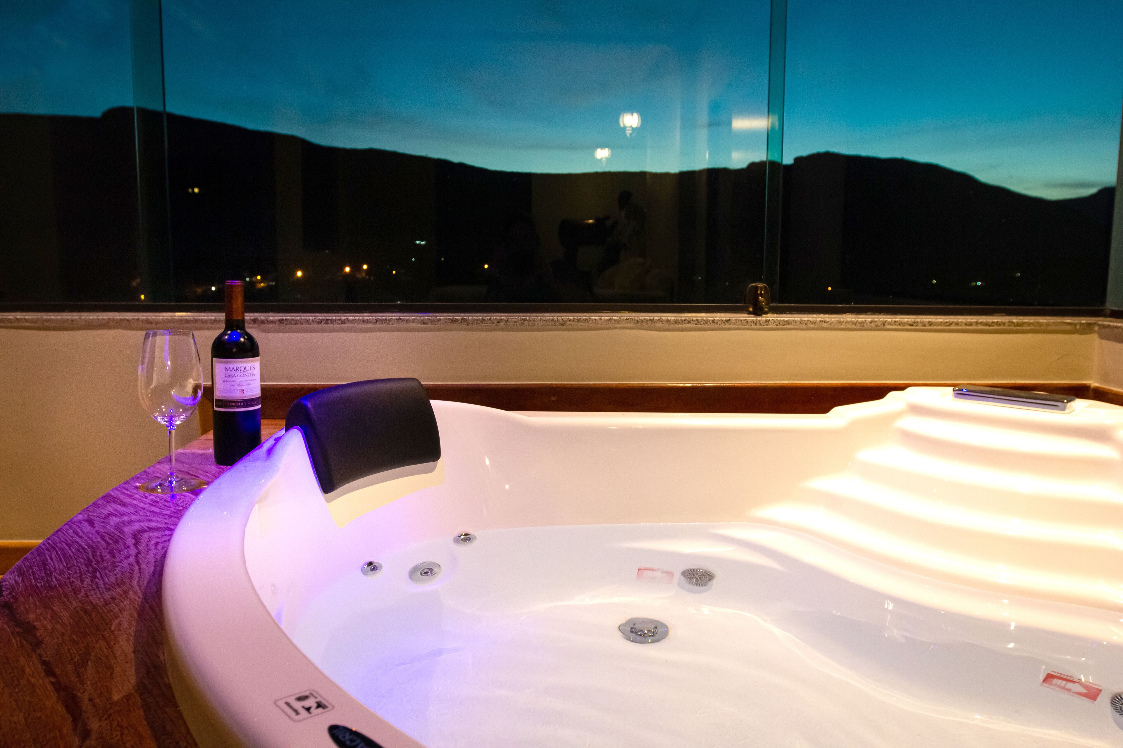 A hot tub with a bottle of wine and a glass on a wooden table, with a view of a cityscape at dusk.