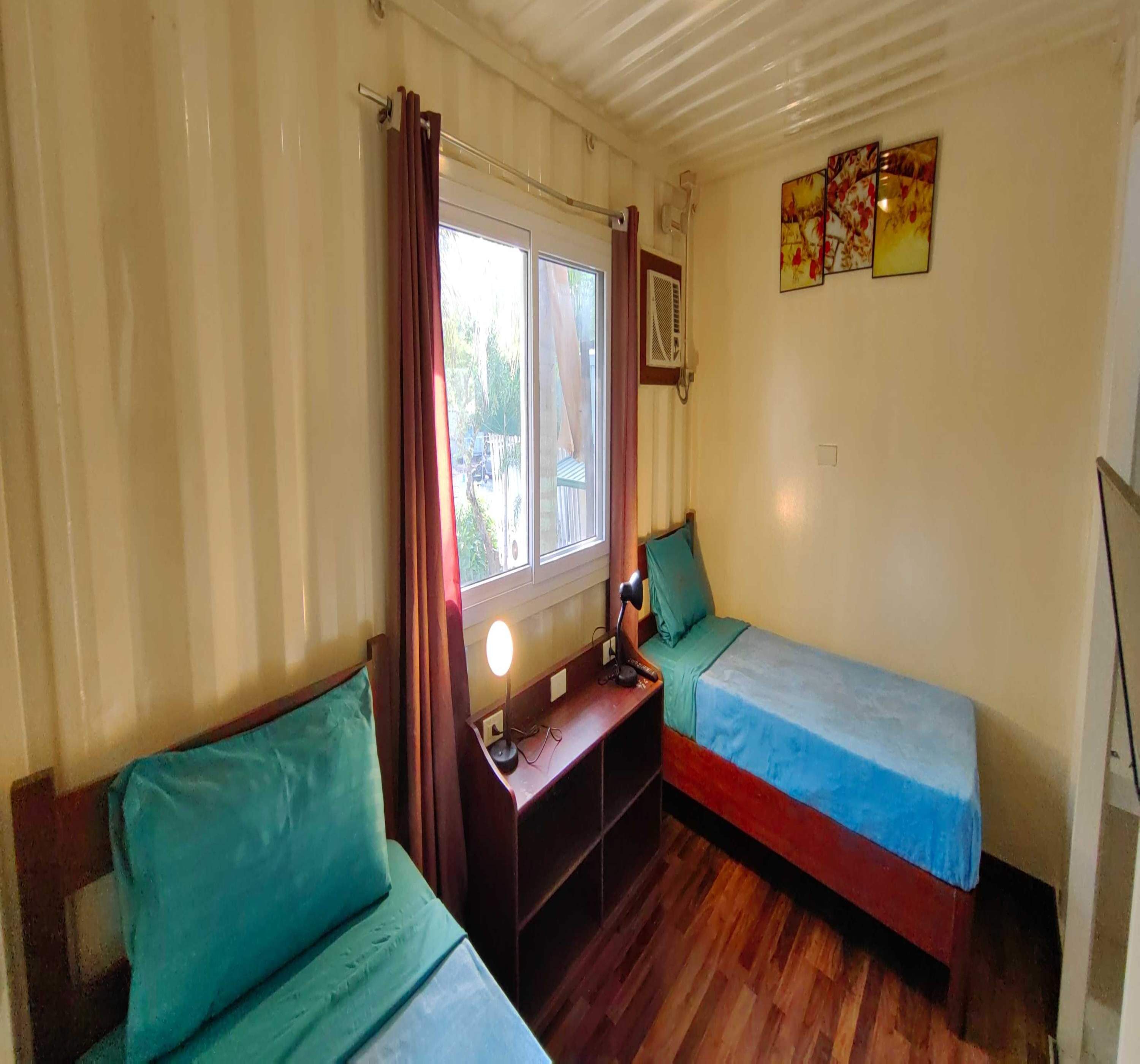 A compact room with two single beds, a small desk with a lamp, a window with curtains, and a wall-mounted air conditioning unit.