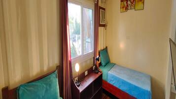 A compact room with two single beds, a small desk with a lamp, a window with curtains, and a wall-mounted air conditioning unit.