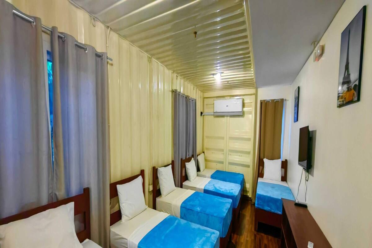 Habitación de hotel con cuatro camas dispuestas en fila, un escritorio de madera, un televisor y un equipo de aire acondicionado en la pared.