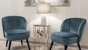 Deux fauteuils de couleur bleu sarcelle avec des pieds noirs, une lampe blanche dotée d’un abat-jour texturé et une table avec un cadre en fil métallique doré.