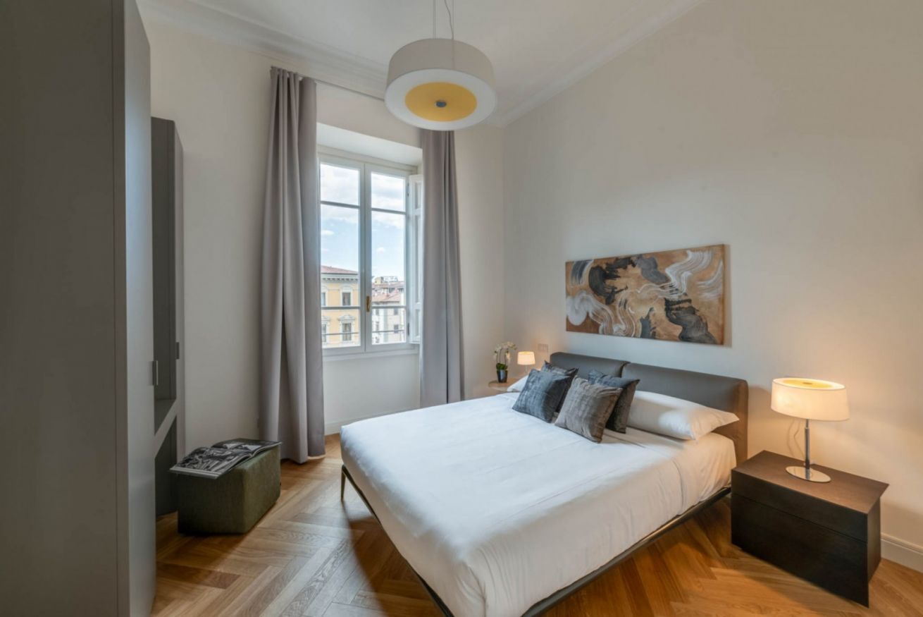 A modern bedroom with a large bed, a bedside table with a lamp, a painting on the wall, and a window with curtains.