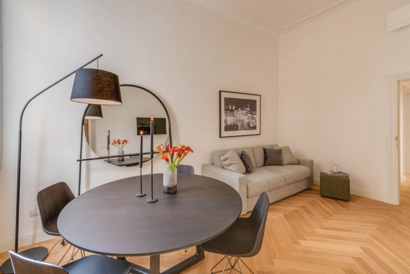 Ein modernes Wohnzimmer mit rundem Tisch, schwarzen Stühlen, einem grauen Sofa, einer Stehlampe, einem Spiegel und einem gerahmten Bild an der Wand.
