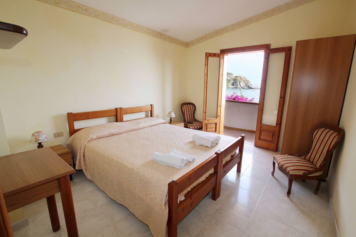A bedroom with a large bed, a wooden desk, a chair, and a view of the sea through an open door.