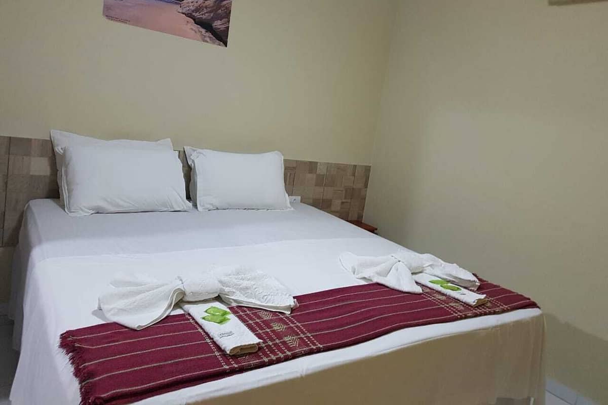Una cama bien tendida con sábanas blancas, una manta a rayas rojas y toallas con diseños de hojas verdes.