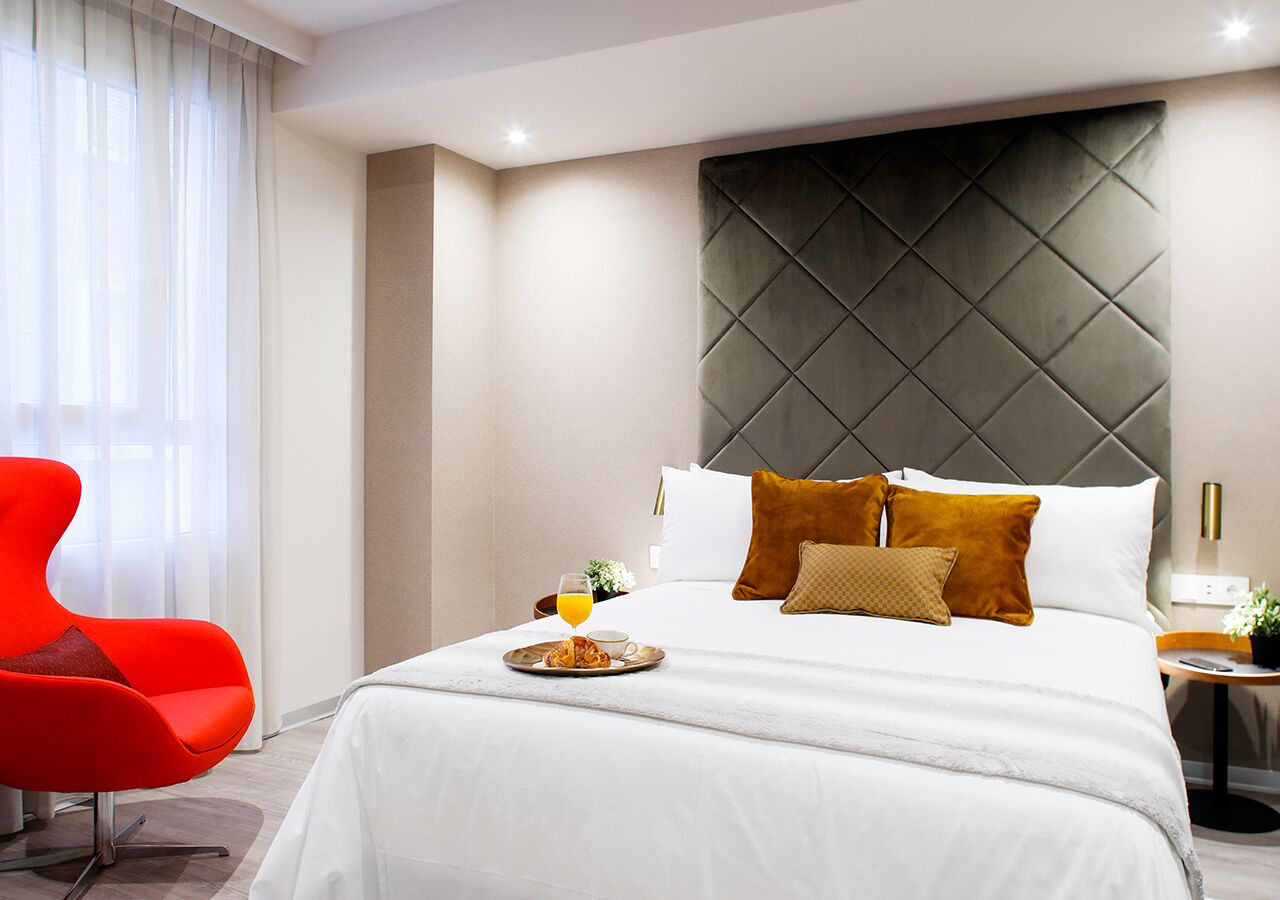 Ett modernt hotellrum med en stor säng, en röd stol och ett brickbord med frukost på sängen.