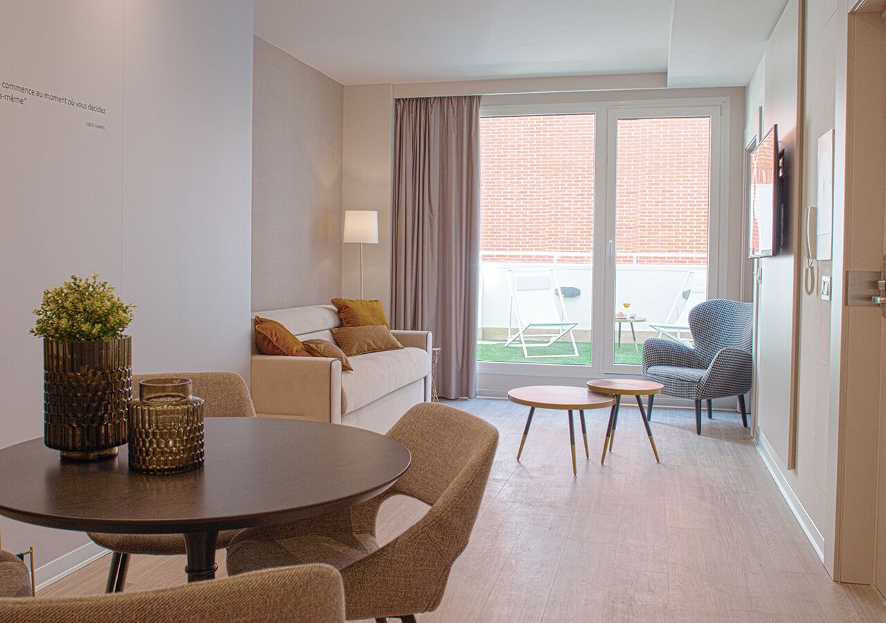 Un salon moderne avec une table à manger ronde, un canapé et un fauteuil. Il y a un balcon avec vue sur un immeuble en briques.